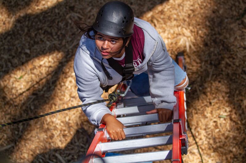 The image shows a person wearing a helmet and safety gear, climbing a ladder. The person appears to be focused and determined. The background is blurred, suggesting an outdoor setting. The ladder is red and silver, and the person is wearing a gray hoodie and jeans.
